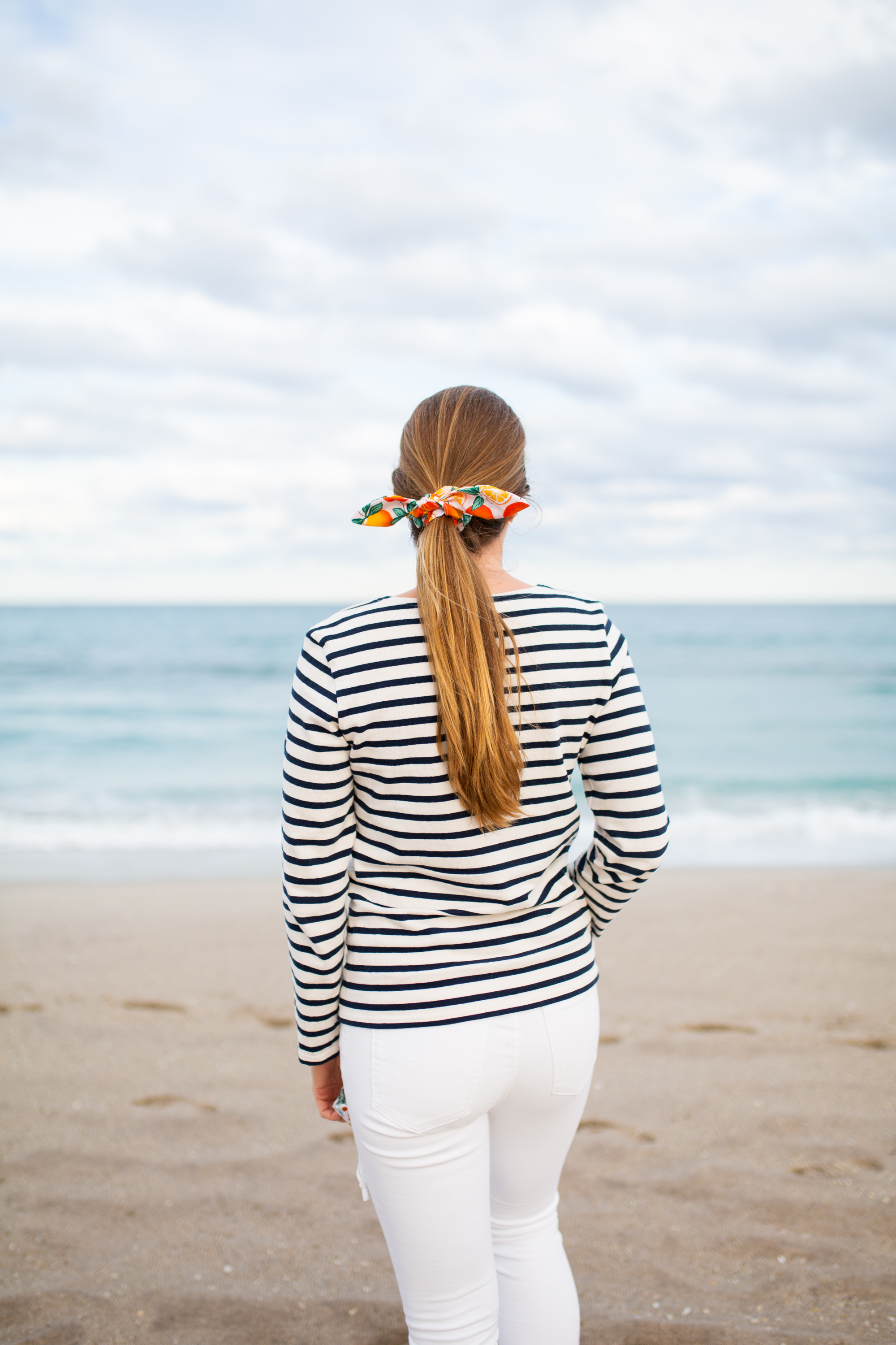 woman standing at the shore wearing striped shirt, white pants, and hair is tied for working full time, running a business, and blogging post