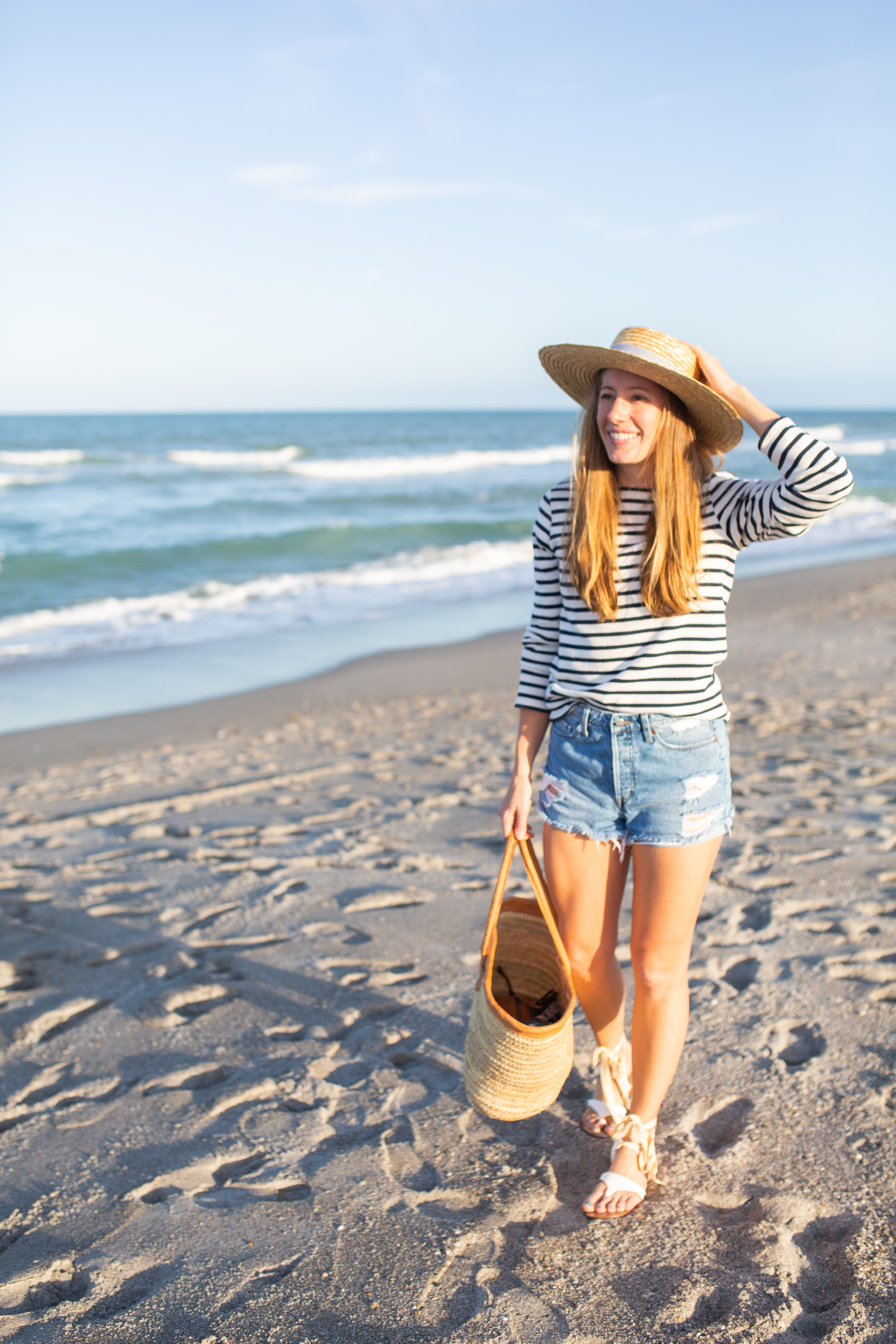 woman wearing denim shorts, long sleeves top, and beach hat and standing at the shore